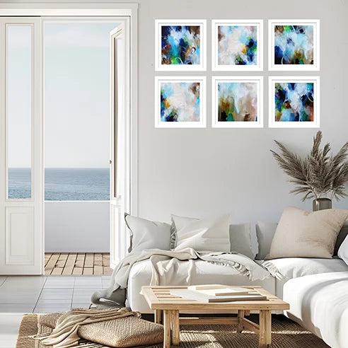Six framed abstract art prints with white frames are arranged in a 2x3 grid on a light gray wall above a white sofa in a bright, modern living room with an ocean view.