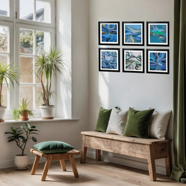 Six framed abstract blue art prints arranged in a 2x3 grid on a white wall above a wooden bench with green and beige pillows, in a bright room with potted plants and a window.