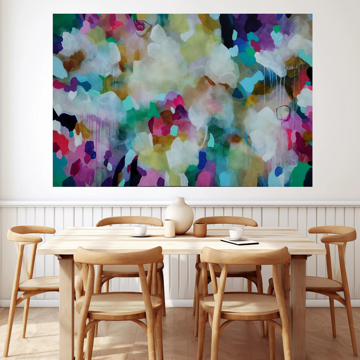 A large abstract canvas wall art print with a vibrant multicolor paint pattern is displayed on a white wall above a light wood dining table and chairs.
