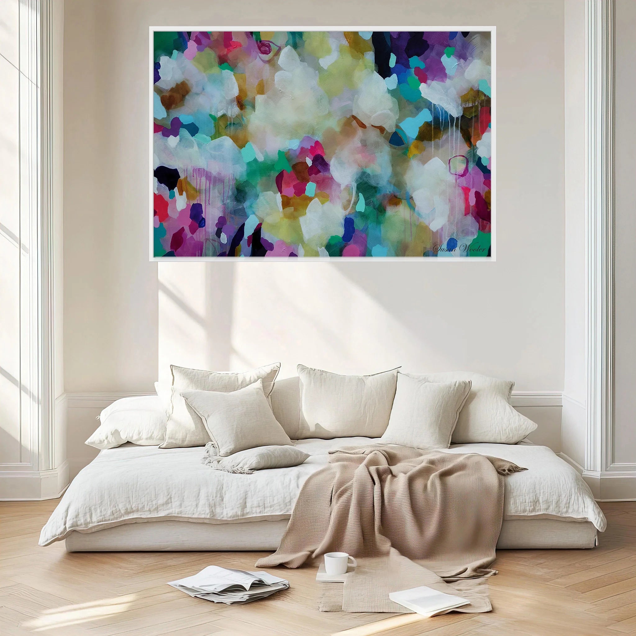 A colorful abstract canvas print with a white frame hangs on a cream wall above a modern low-profile daybed with white linens and a beige throw blanket in a bright, sunlit room.