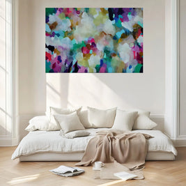 A colorful abstract canvas wall art print featuring a vibrant mix of blue, green, pink, purple, yellow, and white brushstrokes, displayed in a bright, modern living room above a low white sofa with cream pillows and a beige throw blanket.