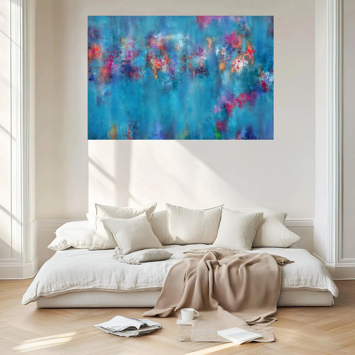 A large abstract canvas print with vibrant blue, purple, and pink brushstrokes hangs on a light beige wall above a modern low white sofa with cream pillows and a beige throw blanket in a bright, sunlit room.