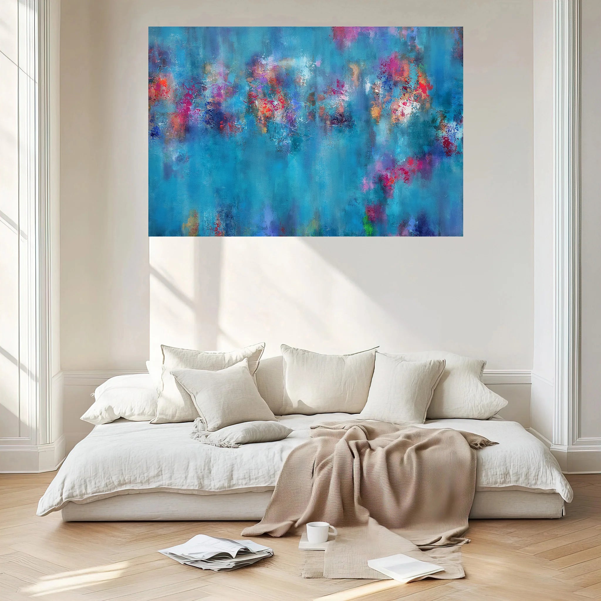 A large abstract canvas print with vibrant blue, purple, and pink brushstrokes hangs on a light beige wall above a modern low white sofa with cream pillows and a beige throw blanket in a bright, sunlit room.