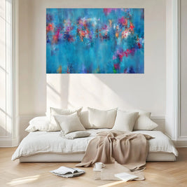 A large abstract canvas print with vibrant blue, purple, and pink brushstrokes hangs on a light beige wall above a modern low white sofa with cream pillows and a beige throw blanket in a bright, sunlit room.