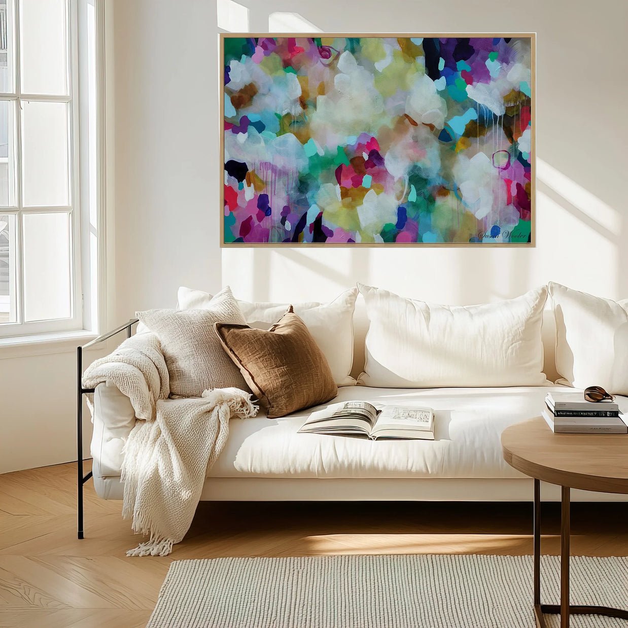 A large framed abstract canvas print with a vibrant multicolor paint pattern hangs on a white wall above a modern white sofa in a bright, sunlit living room.