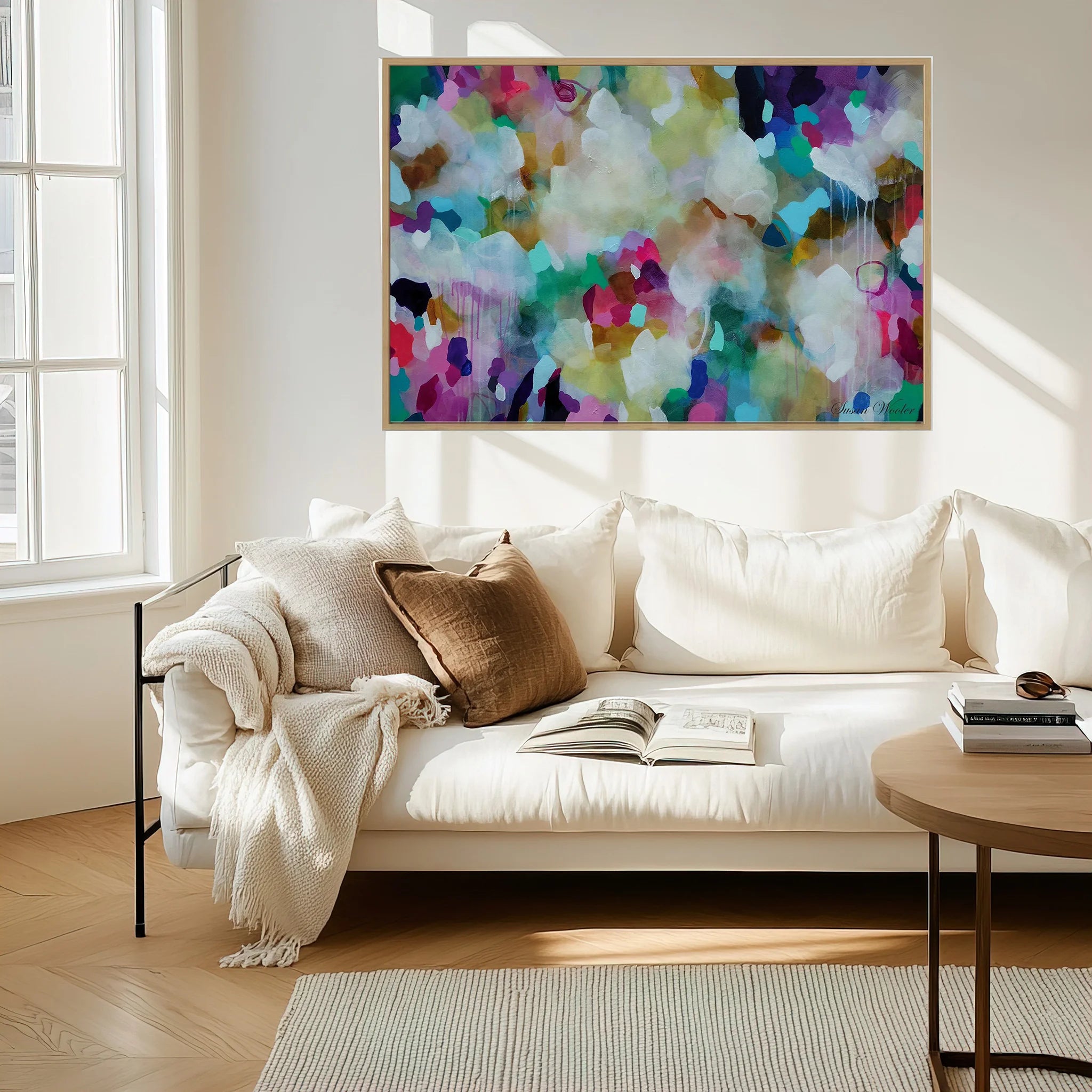 A large framed abstract canvas print with a vibrant multicolor paint pattern hangs on a white wall above a modern white sofa in a bright, sunlit living room.