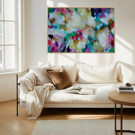 A large framed abstract canvas print with a vibrant multicolor paint pattern hangs on a white wall above a modern white sofa in a bright, sunlit living room.