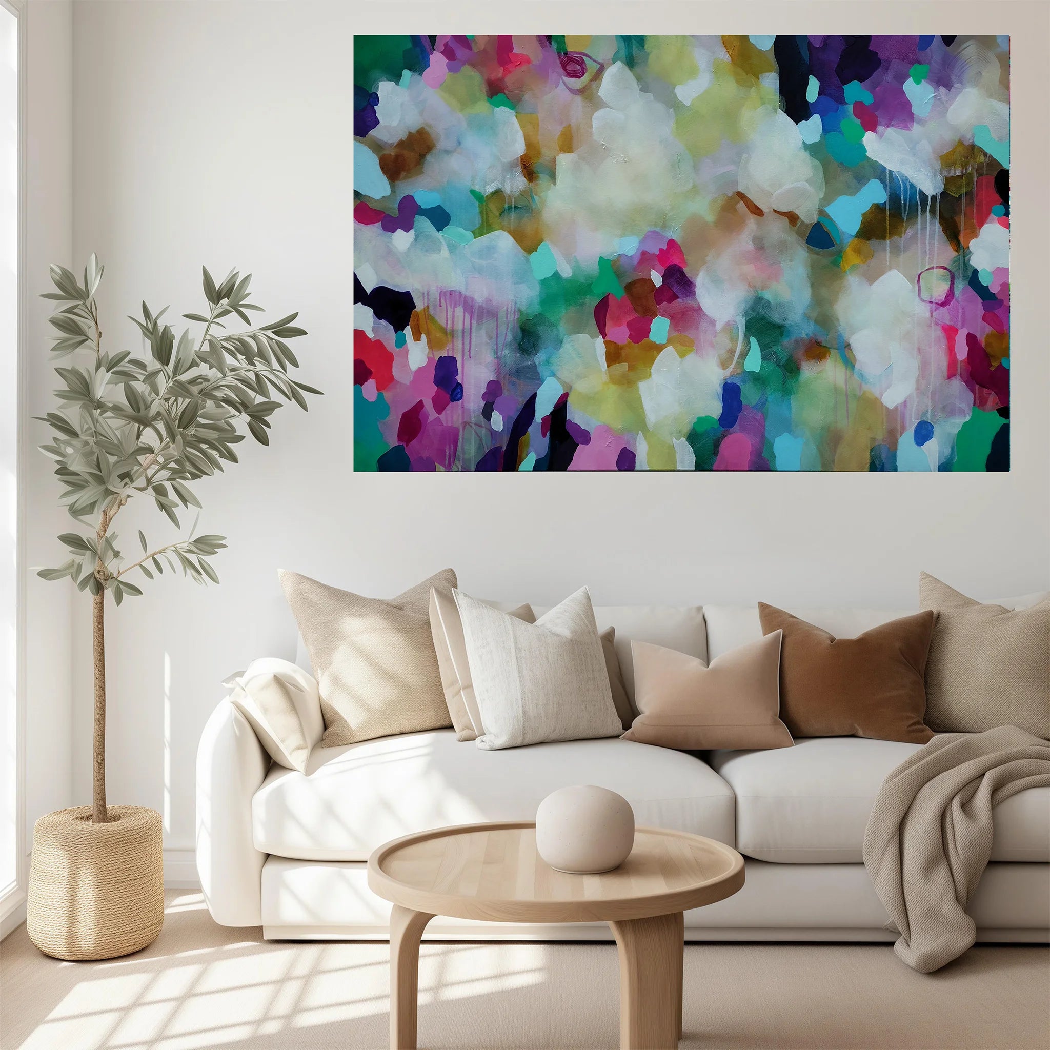 A large abstract canvas print with vibrant multicolor brushstrokes in blue, green, purple, pink, yellow, and white, displayed on a white wall above a modern white sofa with neutral-toned cushions in a bright, contemporary living room.