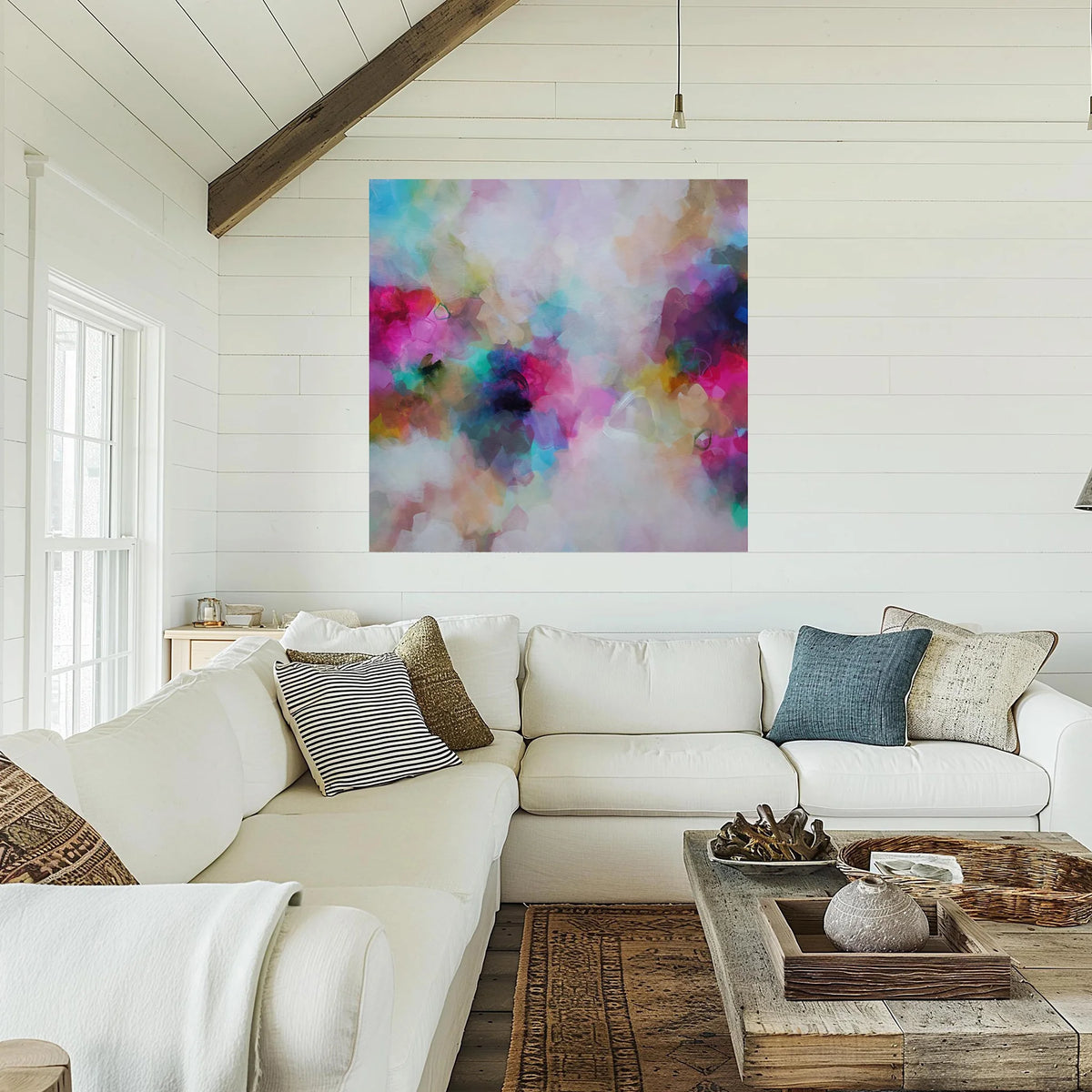 A large square abstract wall art print with a colorful watercolor-like blend of pink, blue, purple, teal, and white hangs on a white shiplap wall above a white sectional sofa in a bright living room.