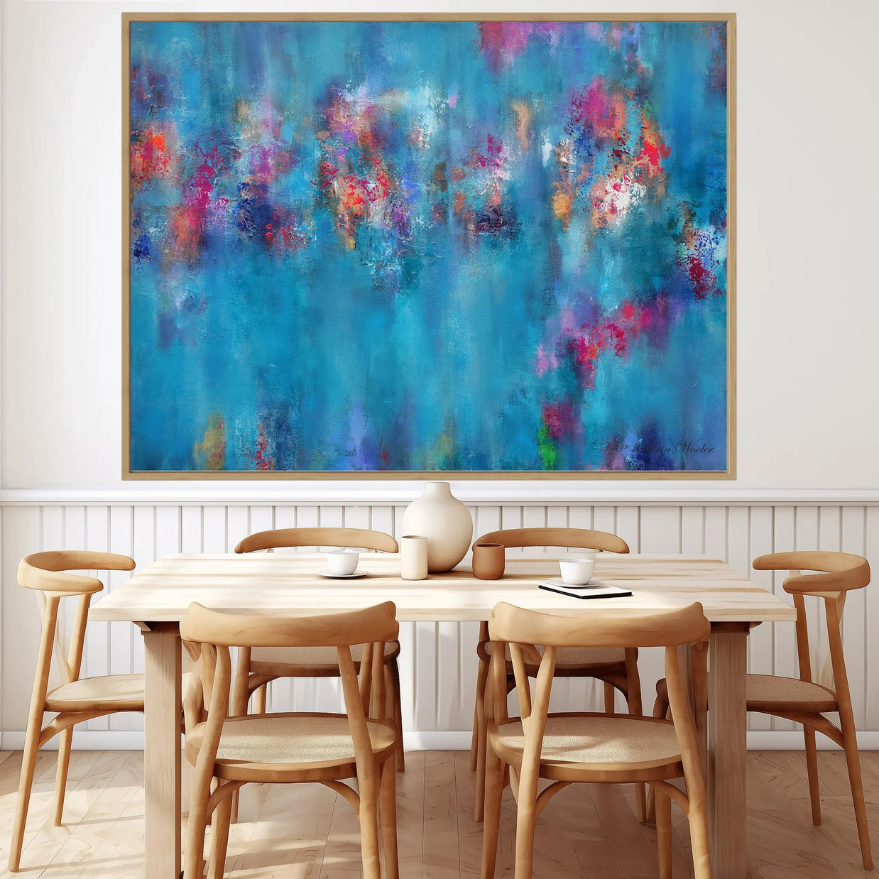 A large framed abstract canvas print featuring vibrant blue, purple, pink, and orange brushstrokes, displayed on a white wall above a light wood dining table and chairs.