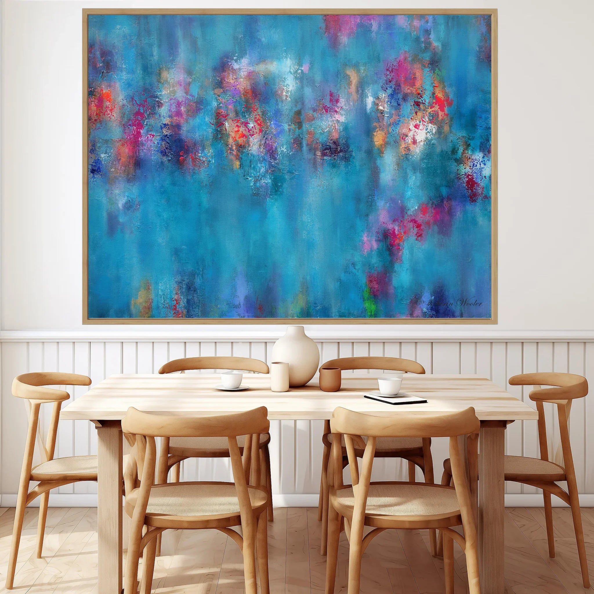 A large framed abstract canvas print featuring vibrant blue, purple, pink, and orange brushstrokes, displayed on a white wall above a light wood dining table and chairs.