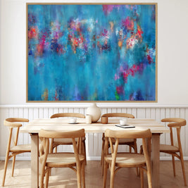 A large framed abstract canvas print featuring vibrant blue, purple, pink, and orange brushstrokes, displayed on a white wall above a light wood dining table and chairs.