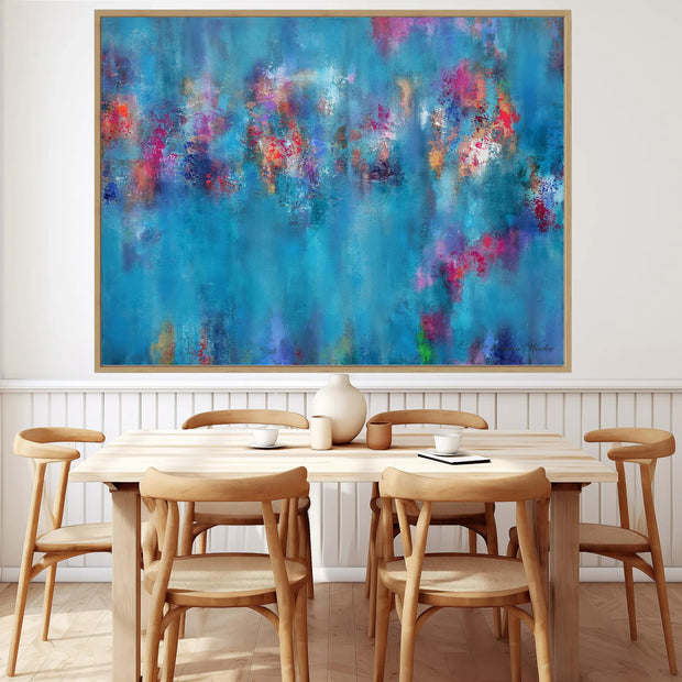 A large framed abstract canvas print featuring vibrant blue, purple, pink, and orange brushstrokes, displayed on a white wall above a light wood dining table and chairs.