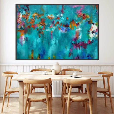 A large framed abstract canvas print featuring a vibrant turquoise, teal, and blue background with splashes of purple, orange, and red, displayed on a white wall above a light wood dining table and chairs.