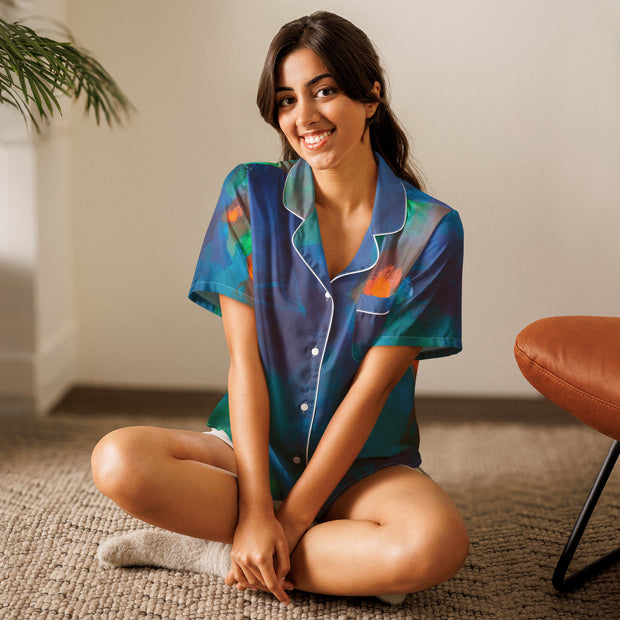 Woman wearing colorful pajama top sitting on the floor with a plant and chair in the background.