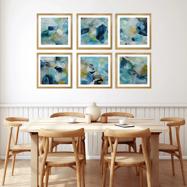Six framed abstract paintings in blue, teal, and white tones with light wood frames are arranged in a 2x3 grid on a white wall above a wooden dining table and chairs.