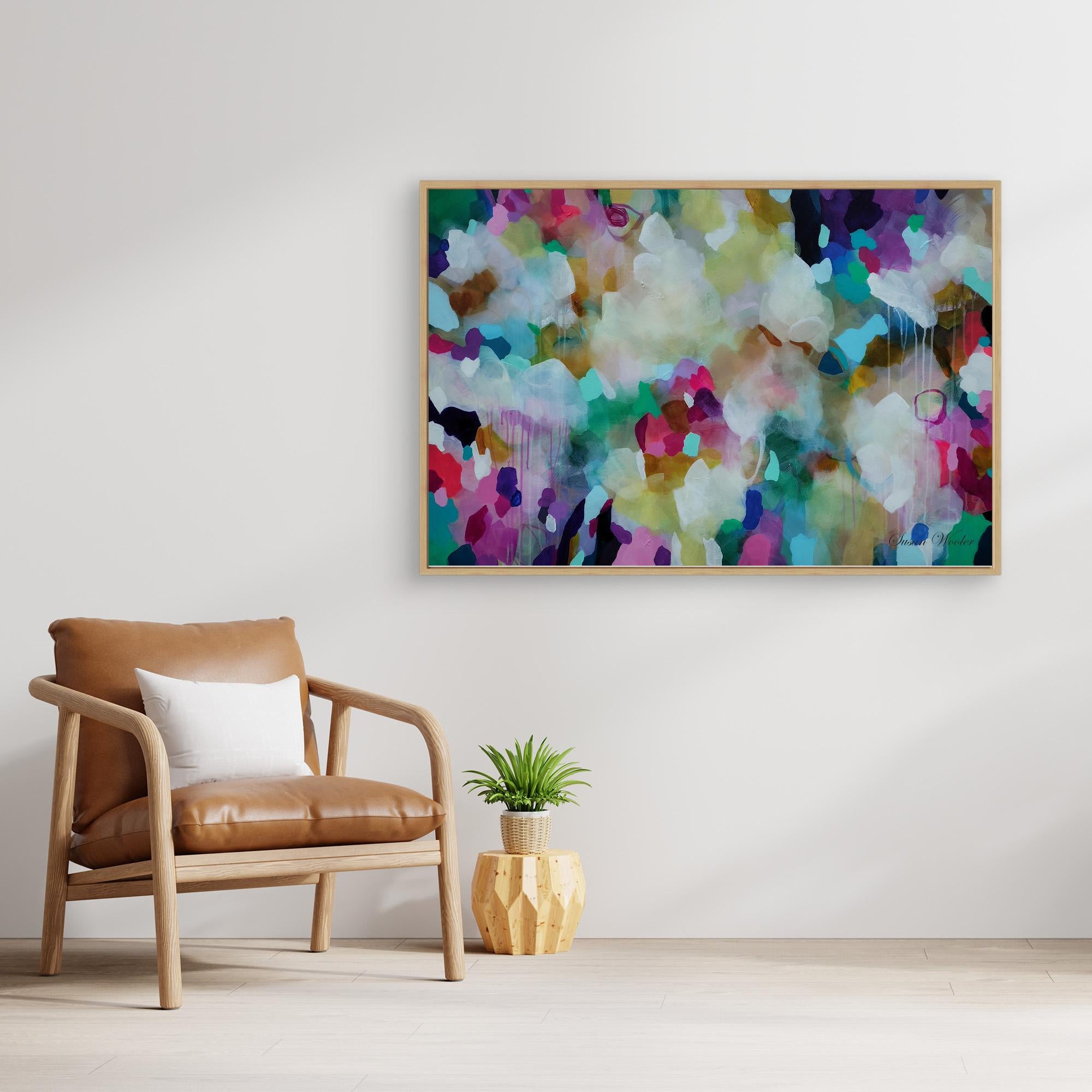 A framed abstract canvas print with a colorful watercolor-like pattern hangs on a light gray wall above a brown leather armchair and a small potted plant.