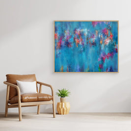 Abstract framed wall art featuring a vibrant blue background with colorful splashes of pink, purple, and red, displayed in a modern living room setting above a brown leather armchair.