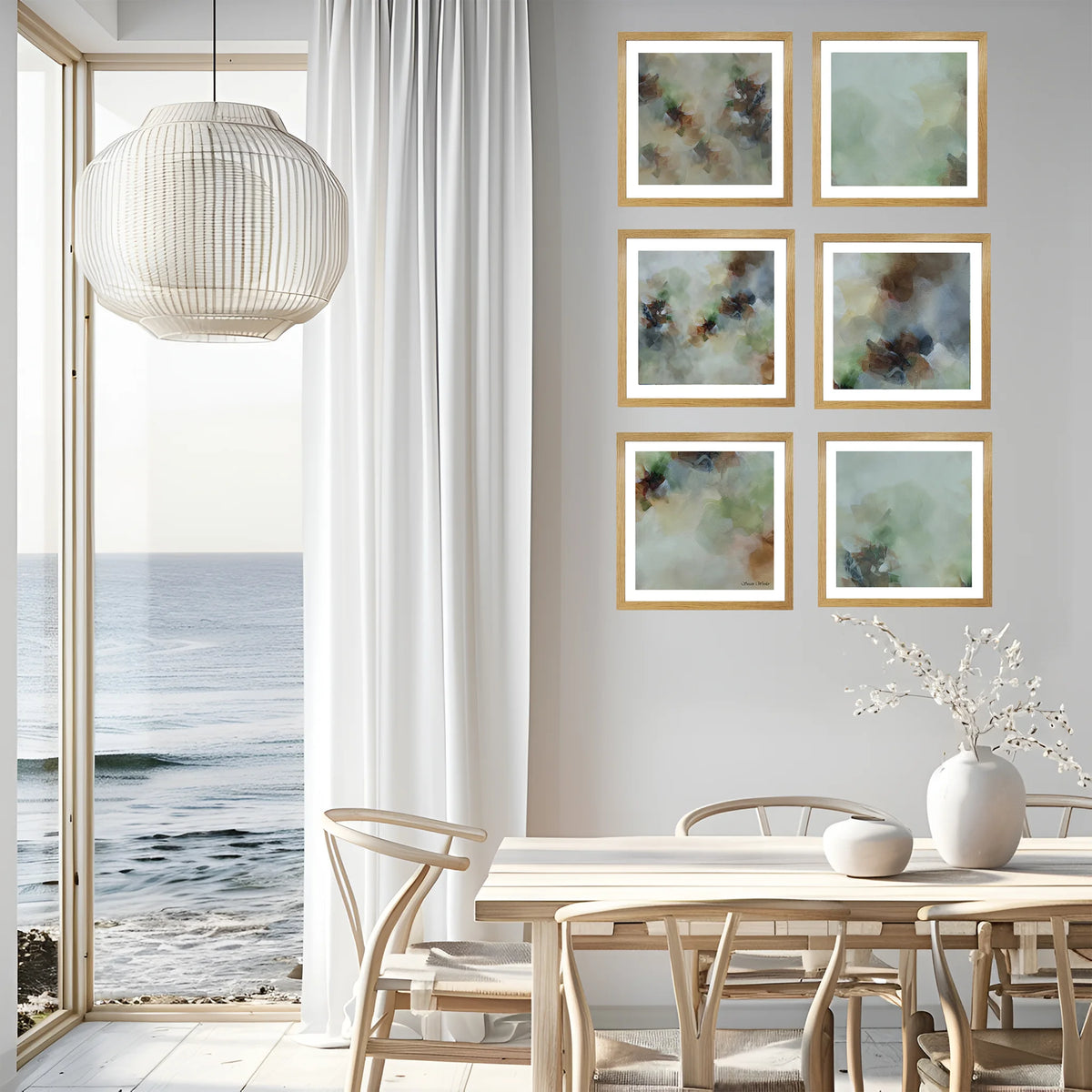 Six framed abstract paintings with soft green, blue, and brown tones, arranged in a 2x3 grid on a light gray wall in a modern dining room with a wooden table, chairs, and a large window overlooking the ocean.