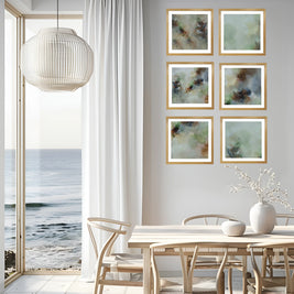 Six framed abstract paintings with soft green, blue, and brown tones, arranged in a 2x3 grid on a light gray wall in a modern dining room with a wooden table, chairs, and a large window overlooking the ocean.