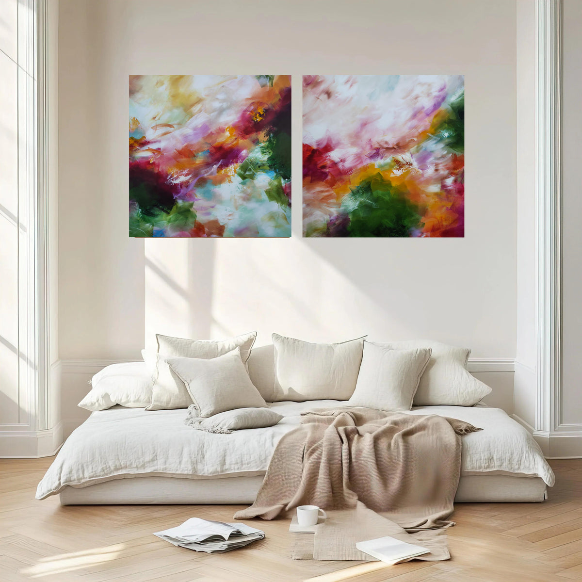 A modern living room setting featuring a two-panel abstract canvas wall art set hanging above a low white sofa with neutral-toned cushions and a beige throw blanket.