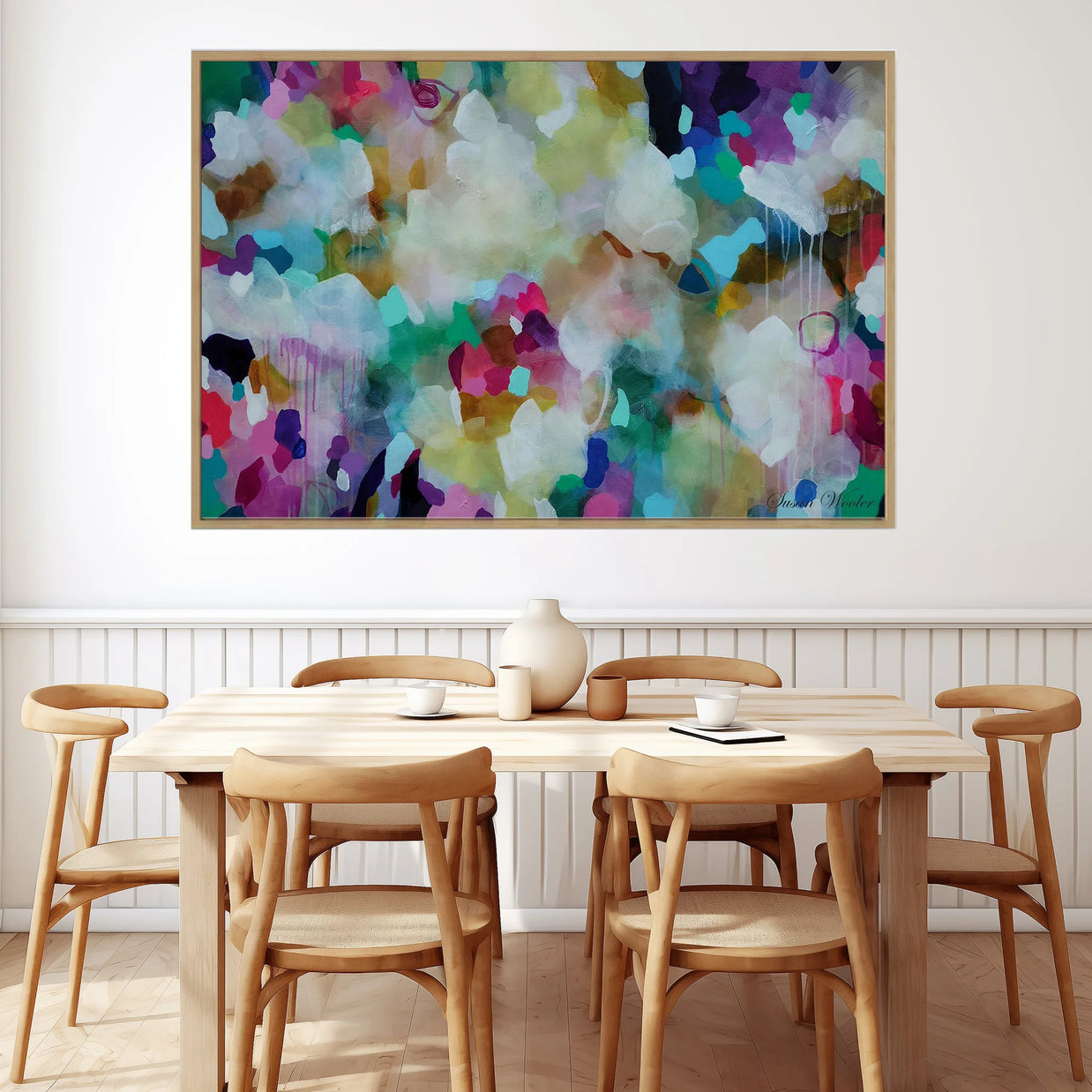 A large framed abstract canvas print with a colorful watercolor-like design in shades of purple, green, blue, white, and yellow, hanging on a white wall above a light wood dining table and chairs.