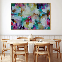 A large framed abstract canvas print with a colorful watercolor-like design in shades of purple, green, blue, white, and yellow, hanging on a white wall above a light wood dining table and chairs.