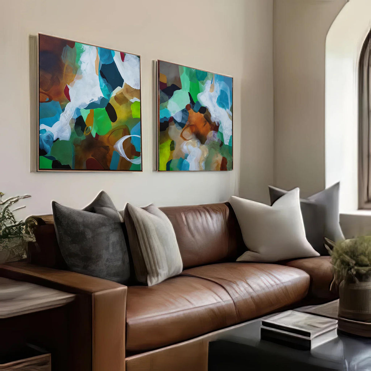 Two framed abstract canvas prints with vibrant green, blue, white, and brown brushstrokes, displayed side-by-side on a beige wall above a brown leather sofa with neutral throw pillows.