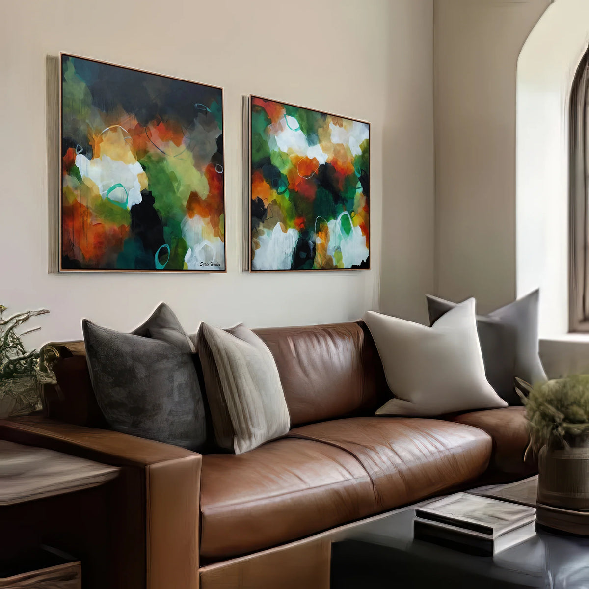 Two framed abstract paintings with green, orange, white, and black brushstrokes hang on a beige wall above a brown leather sofa with gray and cream pillows in a modern living room.