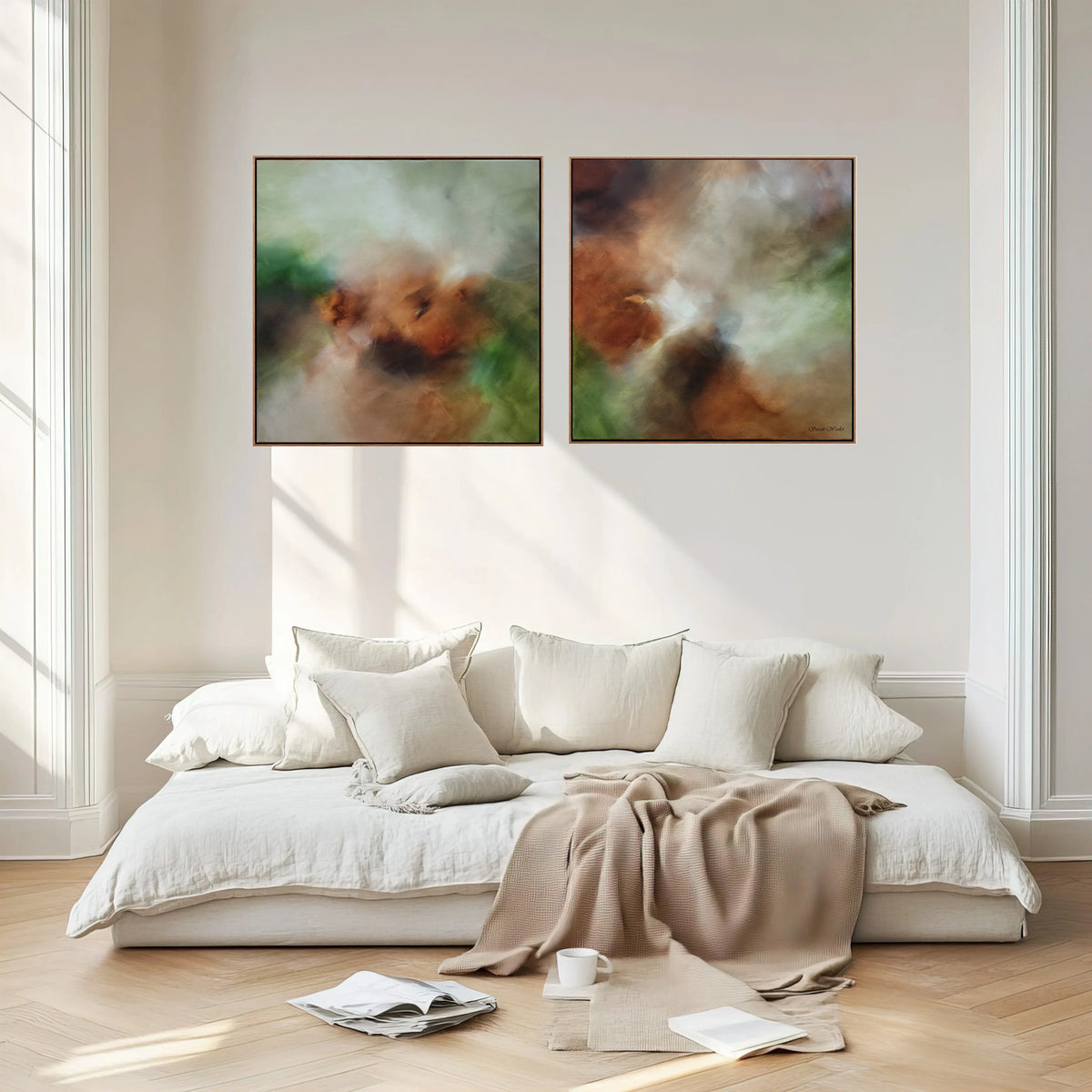 Two abstract framed wall art prints hang above a low white sofa in a bright, neutral room with wood flooring, white bedding, and a draped beige throw blanket.