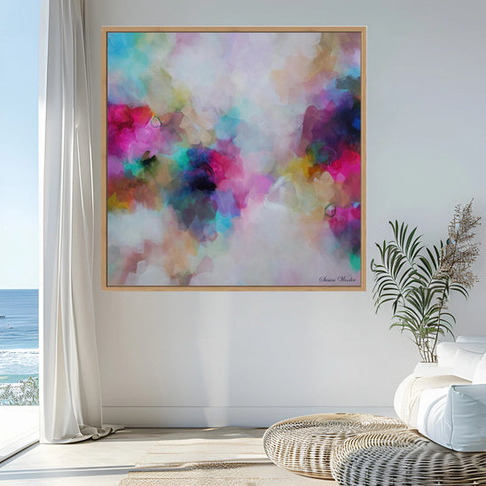 A framed abstract watercolor painting with vibrant pink, blue, purple, green, and yellow tones, hanging on a white wall in a bright modern room with a window view, potted plant, and woven seating.