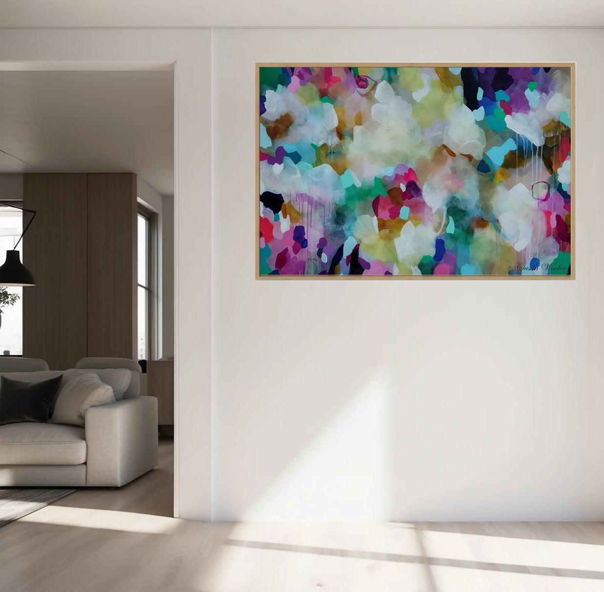 A large framed abstract canvas print with a vibrant multicolor paint pattern hangs on a white wall in a modern, sunlit living room with a light gray sofa visible in the background.