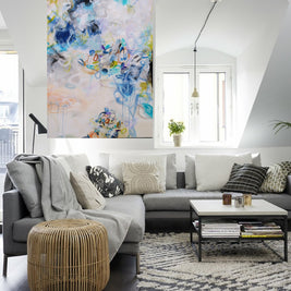 Modern living room with a gray sectional sofa, wicker ottoman, and abstract artwork on the wall.