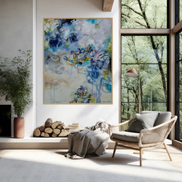 An abstract painting with blue, green, and purple strokes hangs on a white wall in a modern living room with a large window looking out at trees.